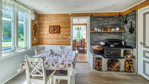 Une cuisine chaleureuse aux murs en bois, une table à manger recouverte d’une nappe à motifs floraux et une cheminée en pierre avec des bûches.