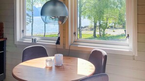 A dining area with a round wooden table, two chairs, and a pendant light. Two windows offer a view of a lake and trees.