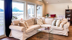 Una sala de estar acogedora con un sofá seccional color beige, una mesa de centro de vidrio y piso de madera. Amplias ventanas ofrecen vistas a un paisaje montañoso.