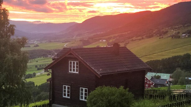 Ein hölzernes Haus mit steilem Dach, umgeben von üppigen grünen Feldern und Hügeln.