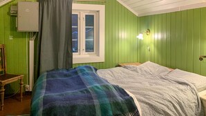 Ein Zimmer mit Bett, einem Fenster mit Vorhängen, einer Nachttischlampe und einem kleinen Tisch.