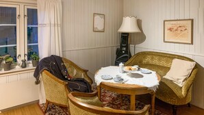 Ein gemütliches Zimmer mit zwei gemusterten Sesseln, einem kleinen runden Tisch mit weißem Tischleinentuch und einer Lampe. Auf einem Fensterbrett stehen Blumentöpfe und an der Wand hängt ein gerahmtes Bild.