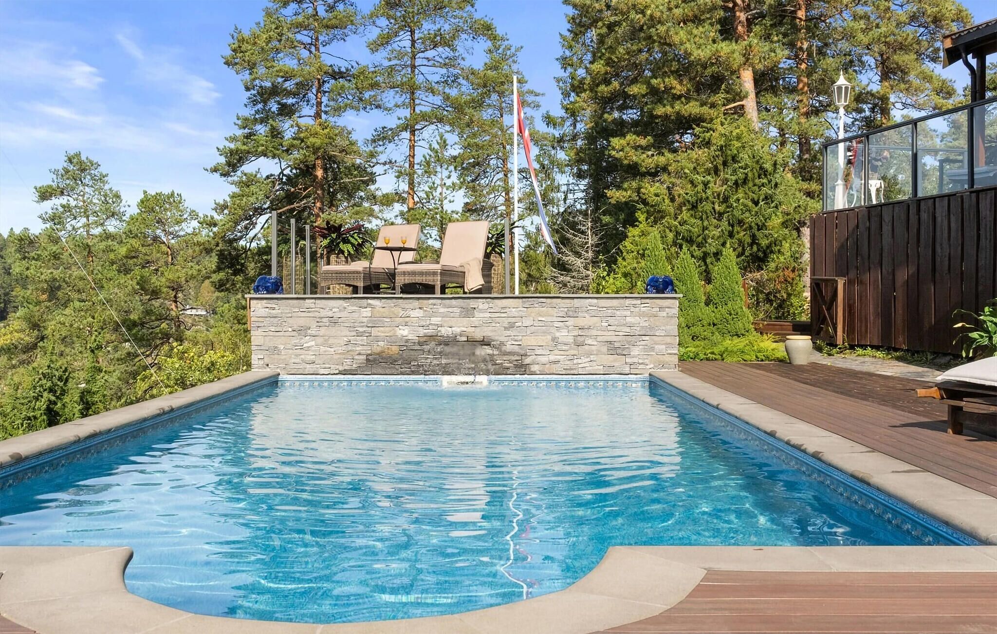 A swimming pool with a stone wall, surrounded by a deck and lounge chairs, with trees and a flag in the background.