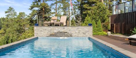 A swimming pool with a stone wall, surrounded by a deck and lounge chairs, with trees and a flag in the background.
