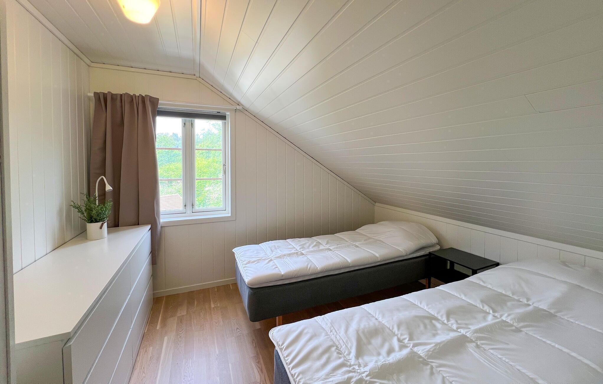 Een kleine kamer met schuine daken, twee bedden, een raam met gordijnen, een nachtkastje met een plant en een houten vloer.