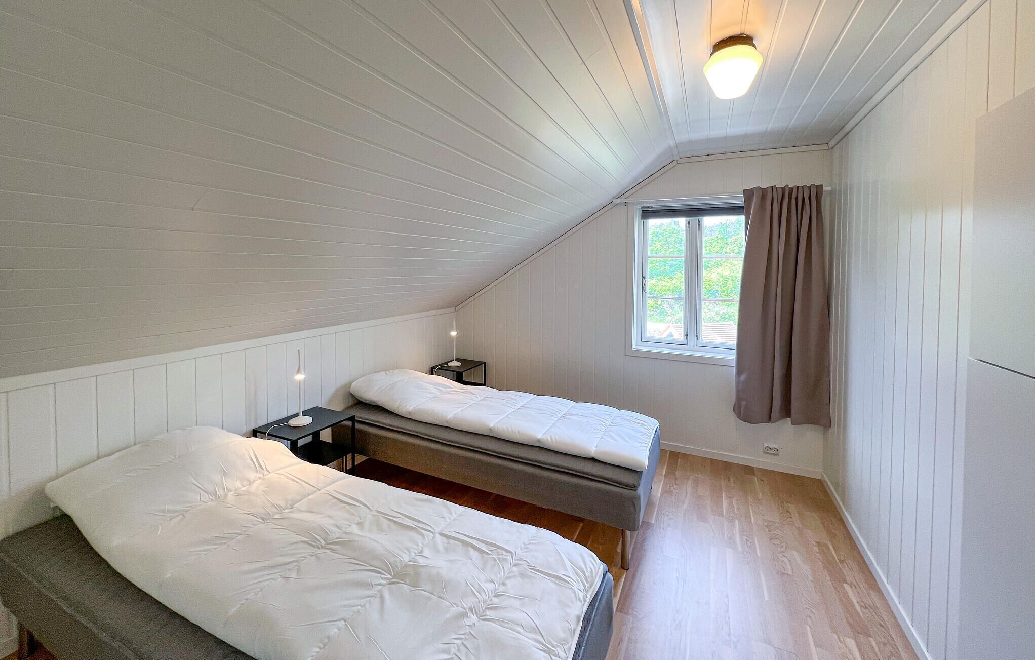 A small, single-room attic bedroom with two beds, a window with curtains, and a ceiling light.