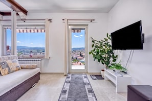 A modern living room with a sofa, a flat-screen TV, a large window with curtains, a potted plant, and a view of a cityscape.