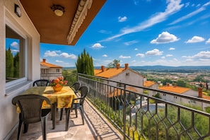 A balcony with a table and chairs, a flower vase, and a view of a village and mountains.