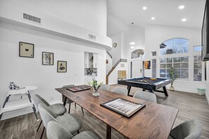 Amplia sala de estar con mesa de comedor de madera, sillas grises, mesa de billar y escalera que lleva a un nivel superior.