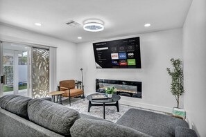 A modern living room with a flat-screen TV mounted on the wall, a comfortable sofa, a small table with decorative items, and a sliding glass door leading to an outdoor area.