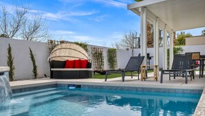 A swimming pool with a built-in waterfall, surrounded by a covered seating area with red cushions, and a patio with lounge chairs.