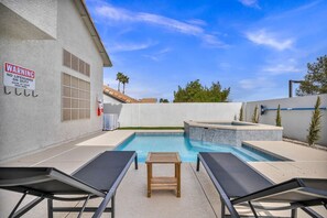 A swimming pool with lounge chairs, a hot tub, and a warning sign on a building.