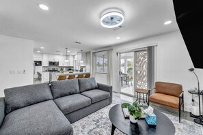 Una sala moderna con un sofá seccional gris, una mesa de centro redonda con una planta y un sillón marrón. Se ve una cocina con armarios blancos y una zona de comedor con mesa y sillas a través de una puerta corrediza de vidrio.