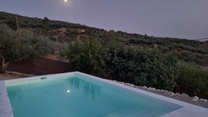 A rectangular outdoor swimming pool with clear water, surrounded by a stone border, set against a backdrop of a hilly landscape with sparse vegetation and a visible moon in the sky. - The Oasis of Periana (Periana)