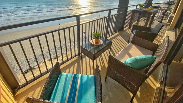 A balcony with wicker furniture, a small table with a plant, and a view of the beach and ocean.