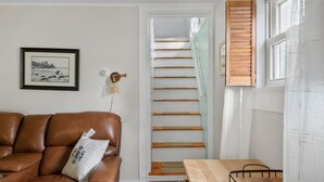 A living room with a brown leather sofa, a framed picture on the wall, a staircase with wooden steps, and a window with blinds. - Adorable Seaside Cottage (Old Orchard Beach)