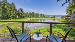 A deck with two chairs and a table overlooking a lake, surrounded by greenery and a cabin. - Waterfront 5-Bedroom Retreat w/ Sauna & Firepit (Juneau)