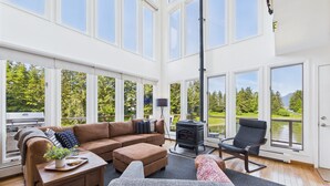 A spacious living room with a large window, a fireplace, and a view of a lake. - Waterfront 5-Bedroom Retreat w/ Sauna & Firepit (Juneau)