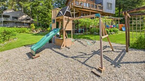 A playground with a slide and swings, surrounded by a wooden playhouse and a house. - Waterfront 5-Bedroom Retreat w/ Sauna & Firepit (Juneau)