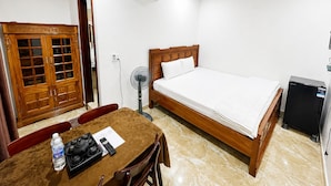 A hotel room with a bed, a desk with a chair, a small refrigerator, an air conditioning unit, and a wooden wardrobe.