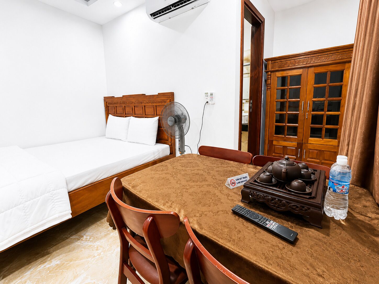Habitación de hotel con cama, una mesa con sillas, un ventilador, una tetera, un control remoto y una botella de agua.