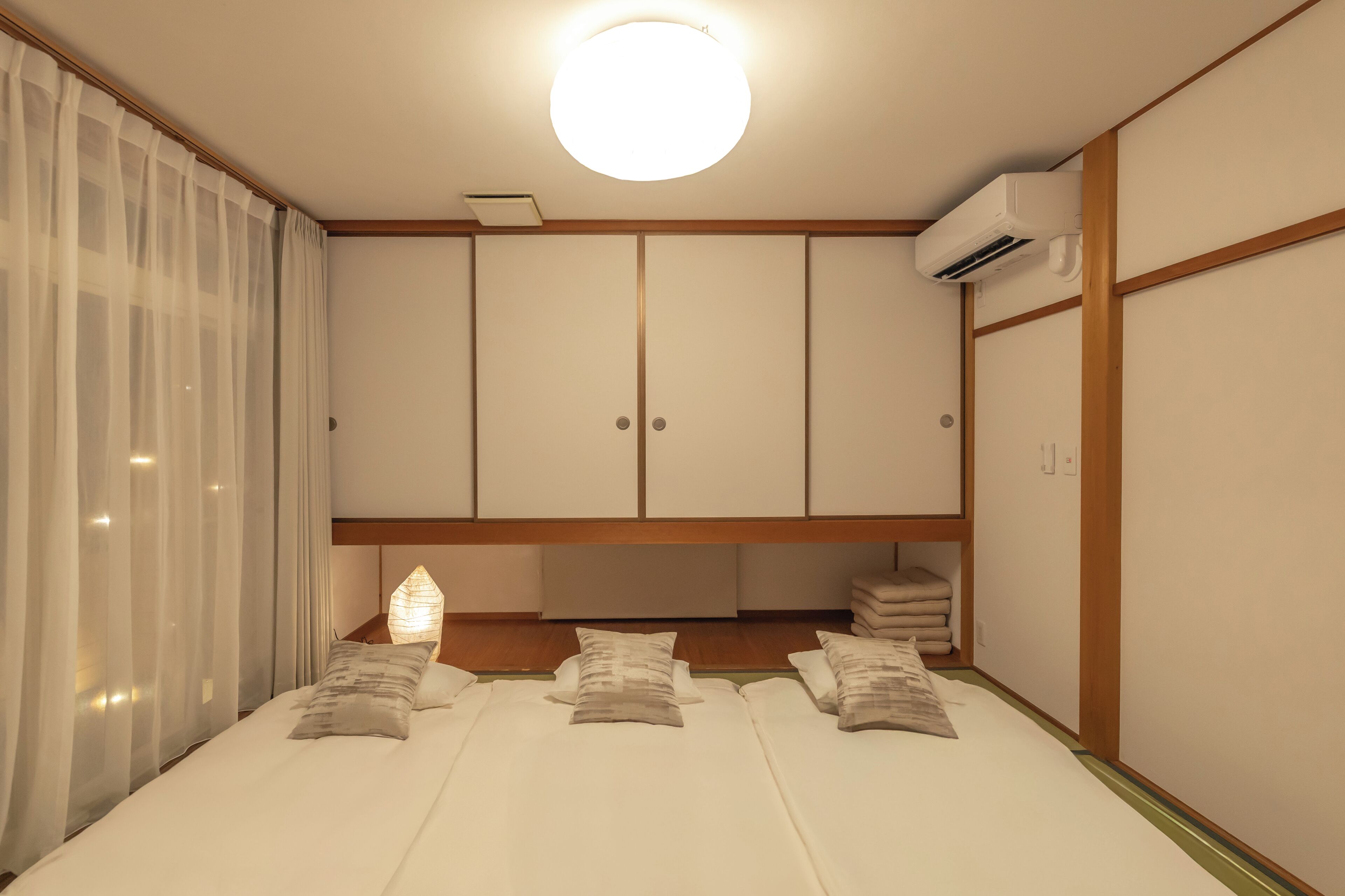 A hotel room with a bed, a bedside table, a nightstand, a wardrobe, and an air conditioning unit.
