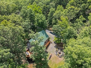 Vista aérea de una cabaña apartada rodeada de un bosque denso, con un edificio principal de techo verde, una terraza de madera y una zona de estar circular.
