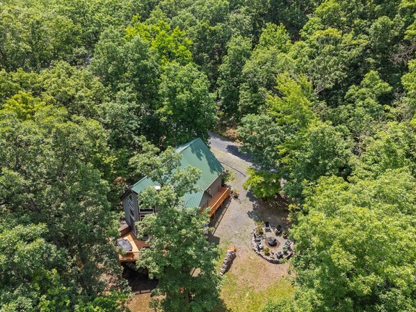 An aerial view of a secluded cabin surrounded by dense forest, featuring a green-roofed main building, a wooden deck, and a circular seating area.