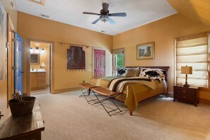 Une chambre à coucher comprenant un lit, un ventilateur de plafond, une table de chevet, une fenêtre avec des stores et une porte donnant sur une salle de bain.