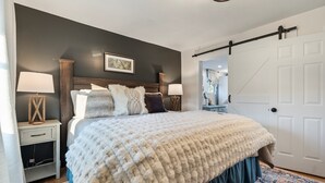 A bedroom with a large bed, two bedside tables with lamps, a dark wall with a framed picture, a white door, and a patterned rug.