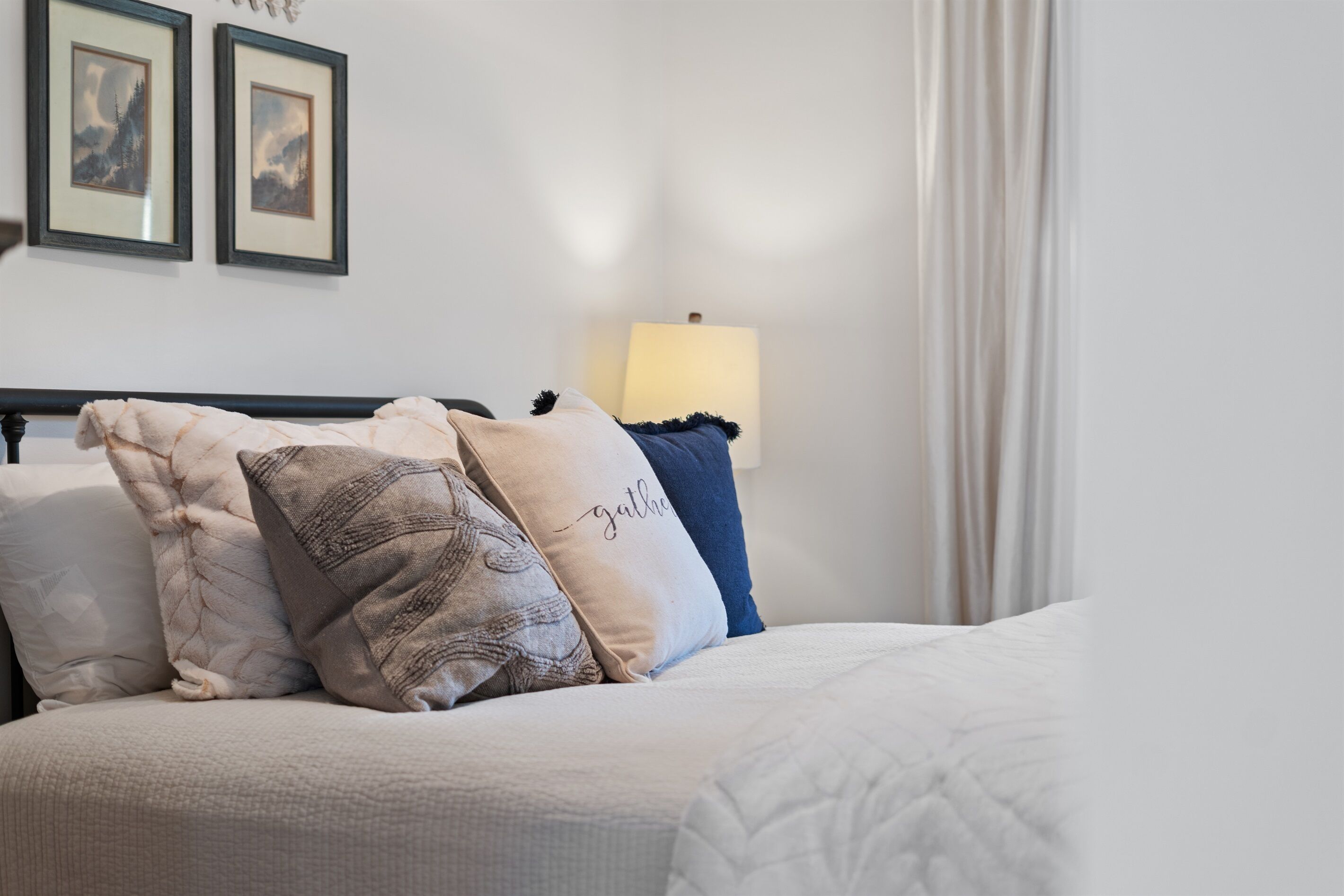 Ein ordentlich bezogenes Bett mit weißer Decke und mehreren Kissen, darunter eines mit der Stickerei "gather". Darüber an der Wand hängen zwei gerahmte Bilder.