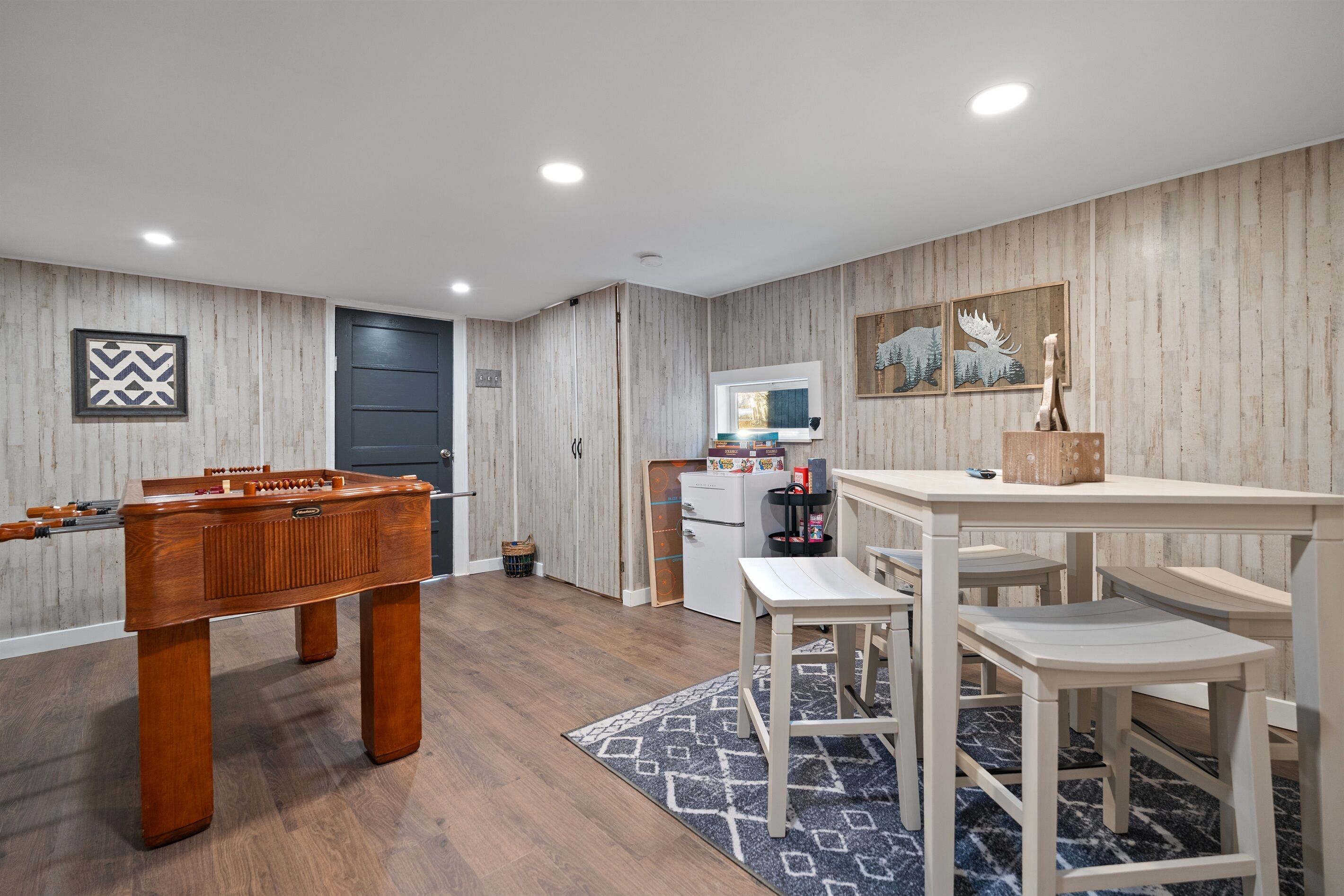 A room with a foosball table, a small kitchenette, a dining area with a table and benches, and a patterned rug.