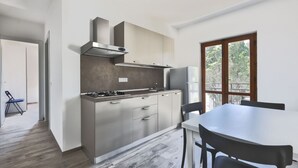 A modern kitchen with a gas stove, stainless steel range hood, and a refrigerator. There is a dining table with chairs and a window with a view of trees. - Bilocale Ida - Goelba (Campo nell'Elba)