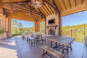An outdoor patio with wooden tables and benches, a stone fireplace, and a TV mounted above it. - Unobstructed Views, Hot Tub, Game Room, Arcade, Family Retreat (Sevierville)