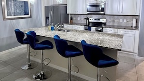 A modern kitchen with white cabinets, a granite countertop, a stainless steel refrigerator, a built-in oven, and three blue bar stools.