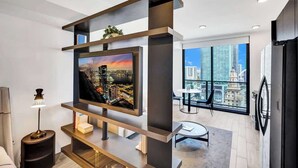 A modern living room with a large flat-screen TV, a dining area with a table and chairs, and a balcony with a city view.