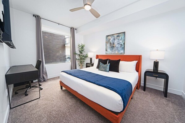 Habitación de hotel con cama, escritorio, silla, mesita de noche, lámpara y ventilador de techo.