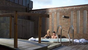 A rooftop pool area with two people relaxing in the water, surrounded by wooden deck and a brick building. - 4 Person Holiday Home on a Holiday Park in Romo (Rømø)