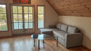 A room with a wooden sloped ceiling, a grey sofa, a glass table with wheels, and a view of a lake through large windows. - Beautiful home in Lepsøy with WiFi (Lepsøy)