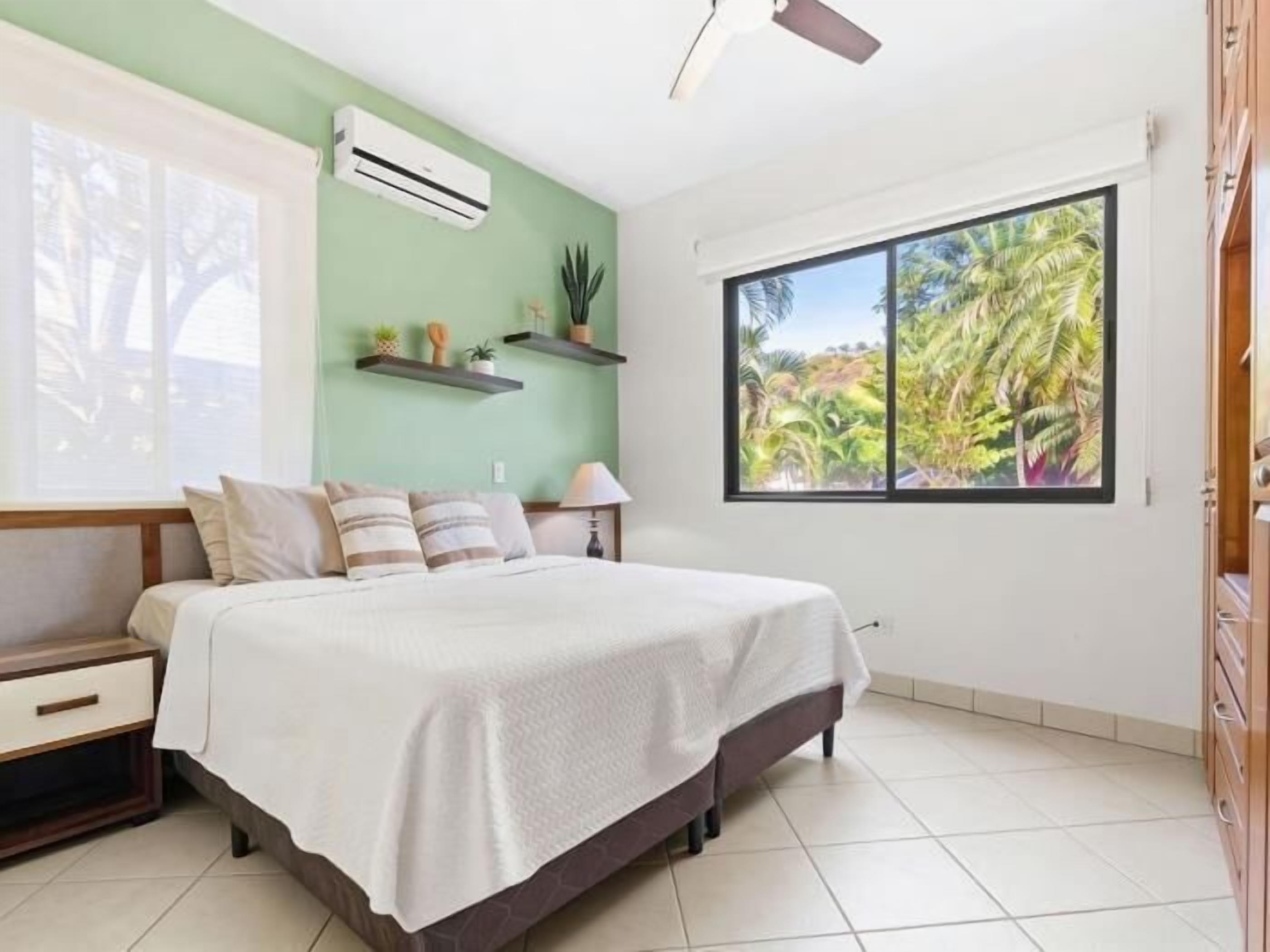 A bedroom with a bed, a nightstand, a dresser, a ceiling fan, an air conditioner, a window with a view of palm trees, and a wall-mounted shelf with decorative items.