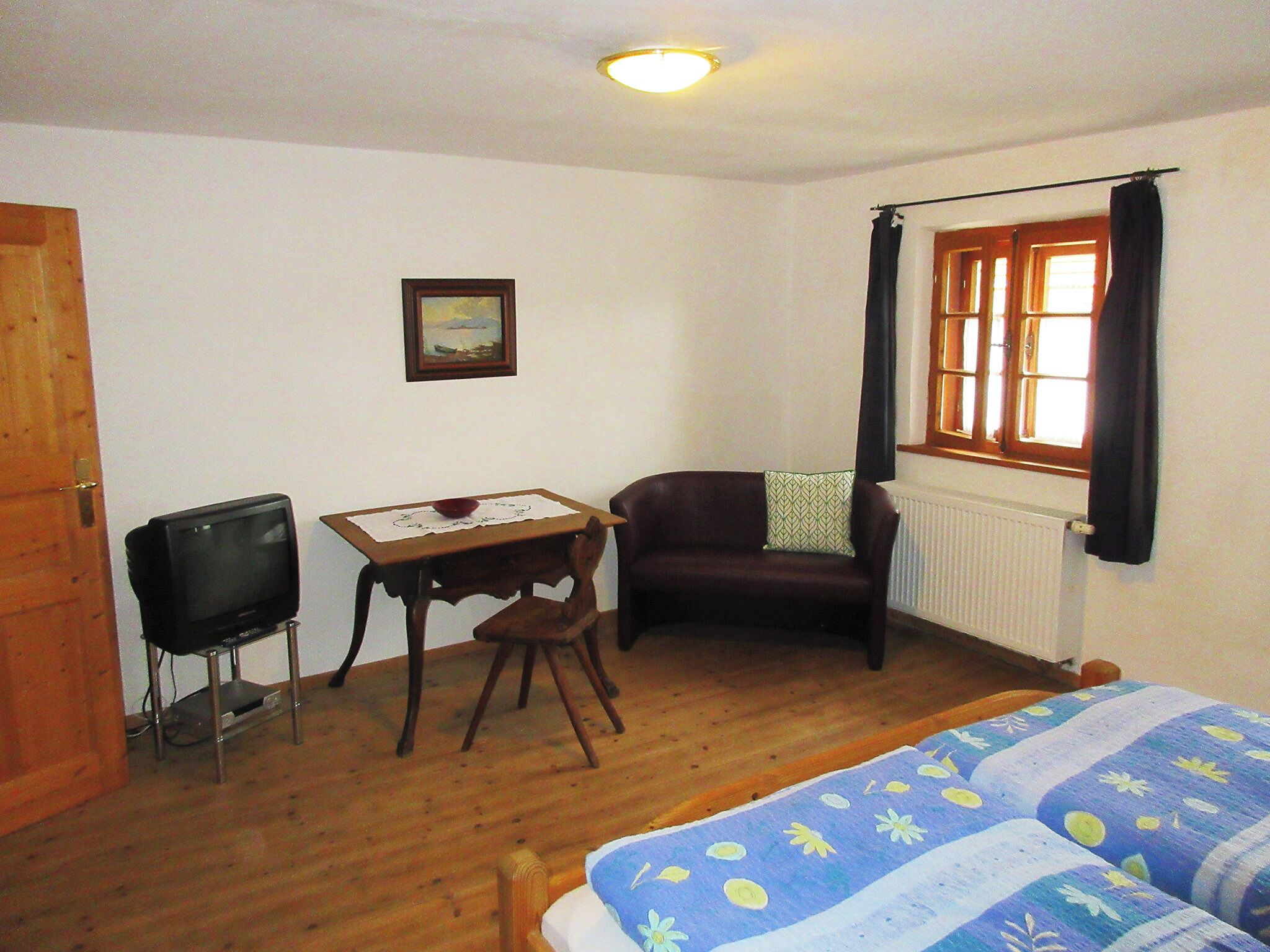 Quarto com cama, escrivaninha, cadeira, televisão, janela com cortinas e radiador.