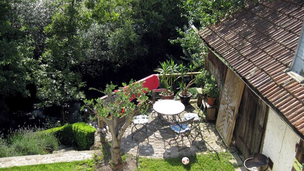 Un jardin avec un espace patio, une petite table et des chaises. On y trouve des plantes en pot et un arbre aux fleurs rouges.