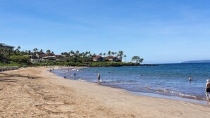 Una playa con cielo despejado, orilla arenosa y algunas personas. Se observan casas con tejados rojos al fondo.