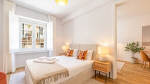 Habitación de hotel moderna con una cama grande, lámparas de noche, una mesita pequeña, una planta en maceta y vistas al balcón exterior.