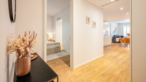Un interior moderno con suelo de madera, un comedor con sillas anaranjadas, y un dormitorio visible con cama y mesita de noche.