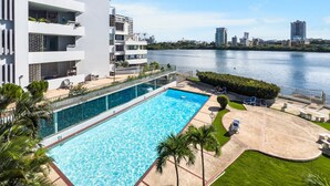 A modern hotel with a swimming pool, a view of the water, and a cityscape in the background. - Condado Studio w/Pool  • Walk to Beach & Dining (San Juan)