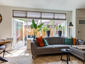 Ett modernt vardagsrum med en grå soffa, ett runt soffbord och en matplats med bord och stolar. Stora fönster med persienner släpper in naturligt ljus och ger utsikt över omgivningen.