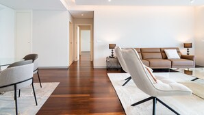 A modern living room with a glass coffee table, a sofa, and two armchairs. There is a wooden floor and a white door in the background.