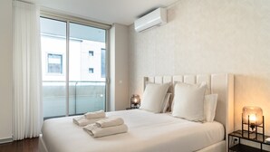 A hotel room with a large bed, white linens, a headboard, bedside lamps, and an air conditioning unit.