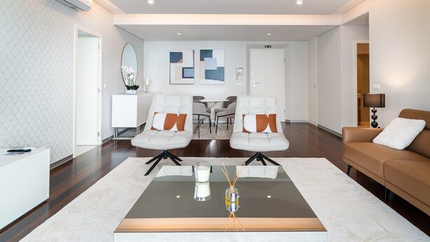 A modern living room with a glass coffee table, two white armchairs with orange cushions, a brown sofa, and a white cabinet.